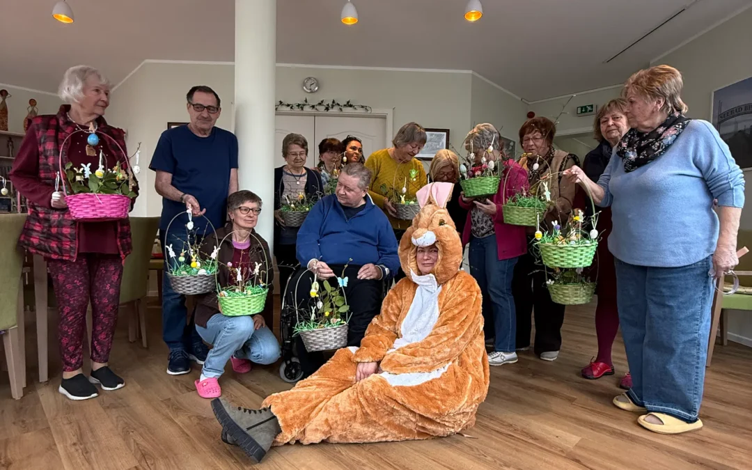 Eine Gruppe strahlender Senioren im betreuten Wohnen hält selbstgebastelte, bunte Osterkörbchen in den Händen, während im Vordergrund ein Vereinsmitglied im flauschigen Osterhasen-Kostüm sitzt.