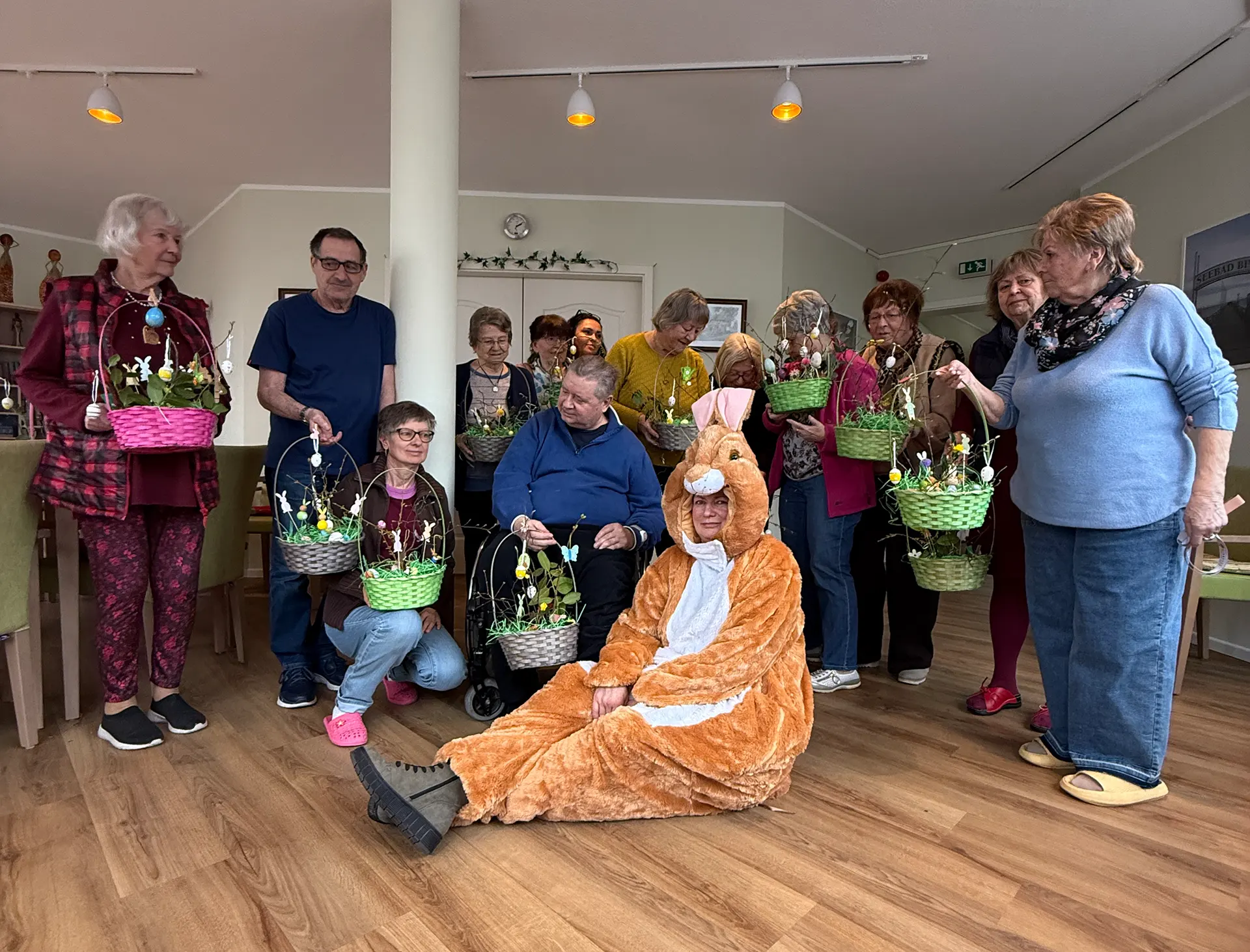 Eine Gruppe strahlender Senioren im betreuten Wohnen hält selbstgebastelte, bunte Osterkörbchen in den Händen, während im Vordergrund ein Vereinsmitglied im flauschigen Osterhasen-Kostüm sitzt.