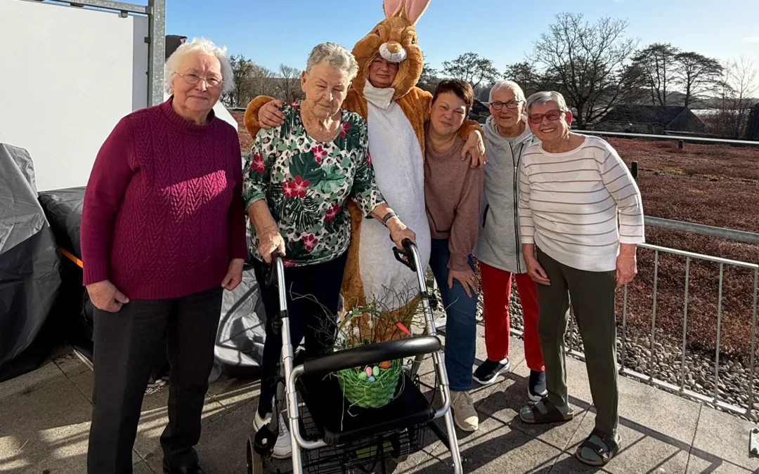 Eine kleine Gruppe strahlender Bewohner des betreuten Wohnens der Volkssolidarität in Binz hält stolz bunte, selbstgebastelte Osterkörbchen in den Händen. Im Vordergrund sitzt ein Vereinsmitglied im lebensgroßen, flauschigen Osterhasen-Kostüm.